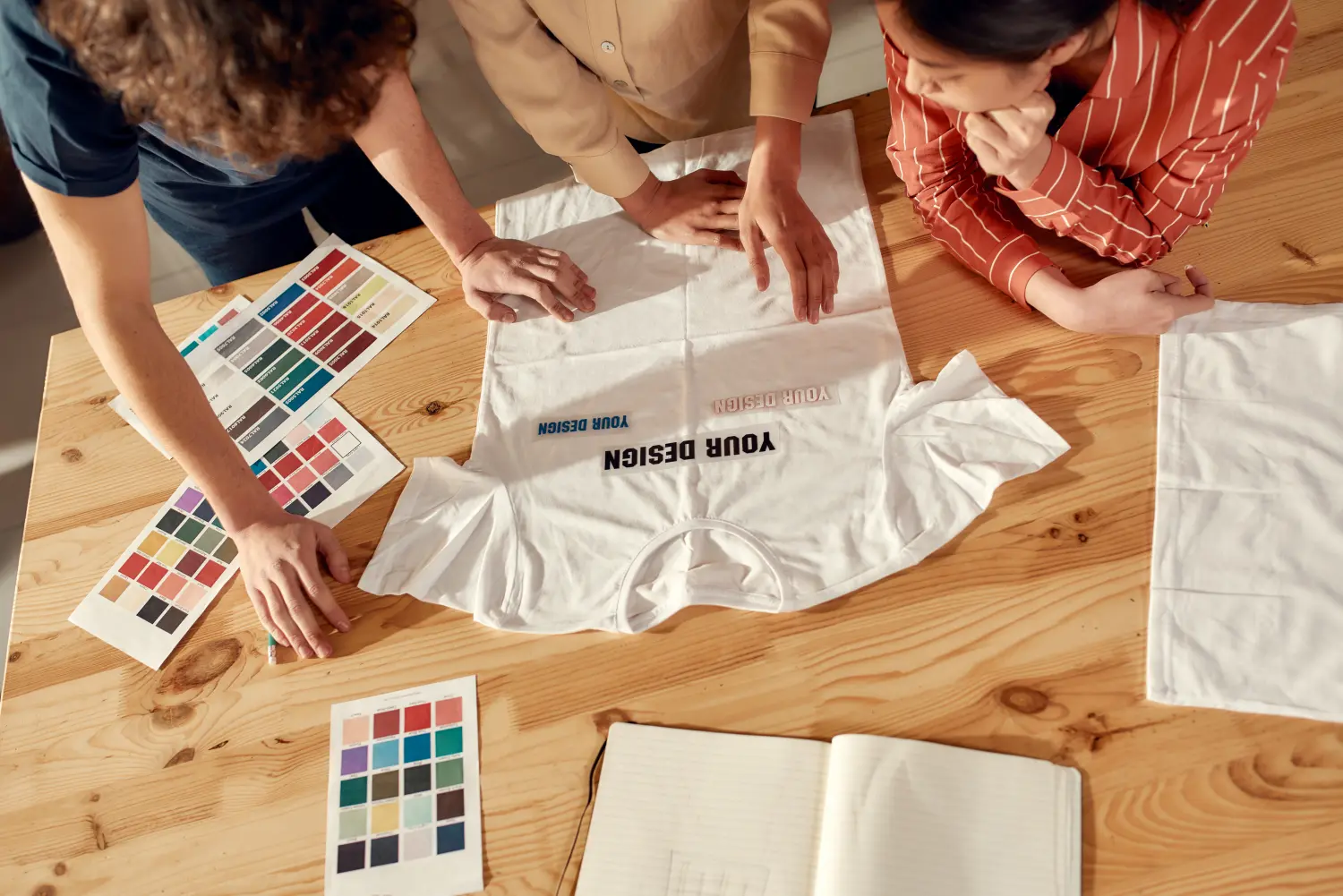 PB Werbeartikel, Textilien mit Druck oder Stick, Schreiben für Ihren Erfolg. Kreative Menschen probieren Aufkleber mit Text an, während sie über Logo und Design von T-Shirts diskutieren. Junger Mann und Frauen arbeiten zusammen in einer Druckerei für individuelle T-Shirts, Kleidung